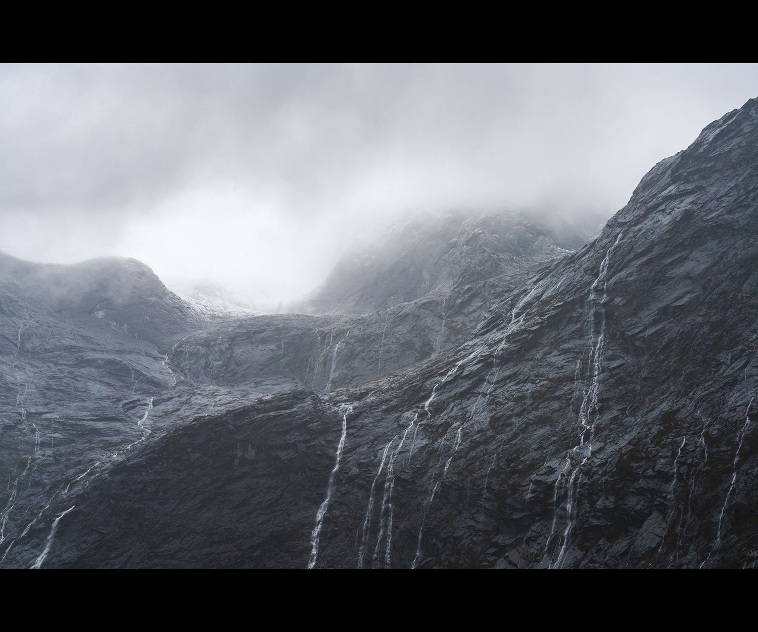 An ominous wall of mountains with waterfalls streaming down. A glacier is barely visible admist the clouds.